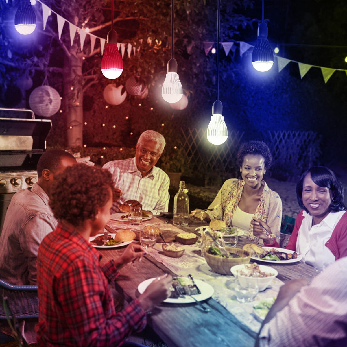 Group of people enjoying a meal outdoors with string lights and a grill in the background.